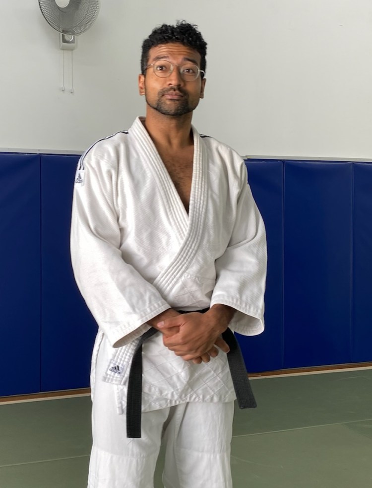A person standing in a judo uniform with a black belt, in a martial arts training room with blue mats and a fan in the background.