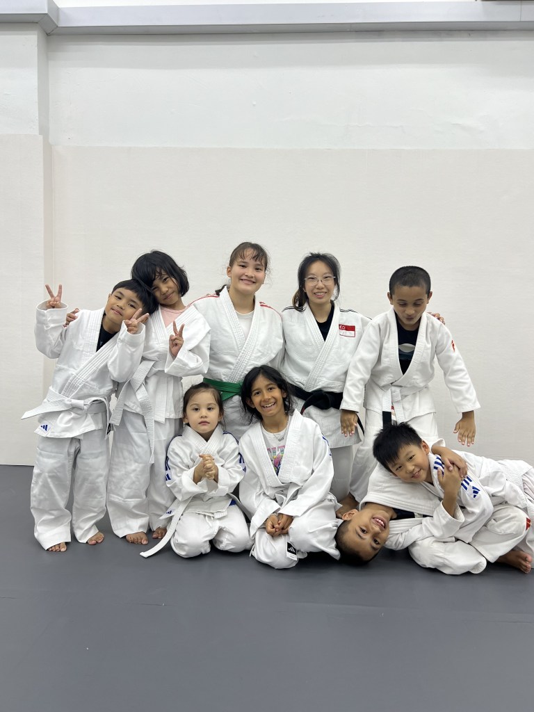 A group of children wearing white judo uniforms posing together on a mat, with some making peace signs and others smiling.