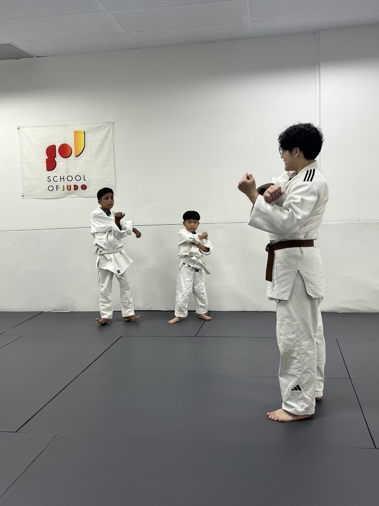 Three children in judo uniforms practicing in a judo dojo with a school banner on the wall.
